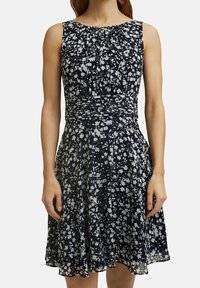 Ärmlös A-linje klänning med marin bakgrund och vitt blommönster, samlad midja, bärs av en stående person med avslappnade armar.