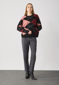 Un pull à motif argyle noir et rose en tissu doux, associé à un pantalon gris slim et des bottes à talons aiguilles noires sur une surface lisse.