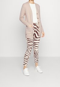 Persona che indossa un cardigan beige con bottoni, una camicia bianca, pantaloni a strisce zebrate marroni e bianchi e sneakers bianche, in piedi contro uno sfondo semplice.