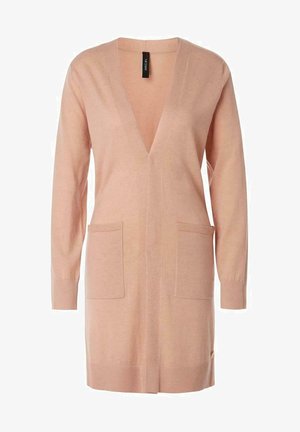 Kvinde iført en beige åben cardigan, mønstret bluse, sort lædernederdel og sorte vævede platformsandaler, stående mod en hvid baggrund.