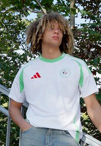 Maglia sportiva bianca con accenti verdi, con il marchio Adidas e un logo circolare. Realizzata in materiale leggero, con maniche corte e colletto girocollo.