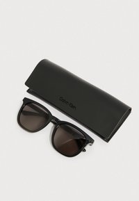 Okulary przeciwsłoneczne Calvin Klein z czarnymi plastikowymi oprawkami i ciemnymi szkłami, wraz z matowym czarnym etui z subtelnym logo.
