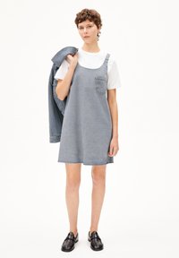 Blauwe denim jurk met brede banden, een voorzak en een rafelige zoom. Gecombineerd met een wit t-shirt en zwarte loafer schoenen. Denimjasje over de arm gedrapeerd.