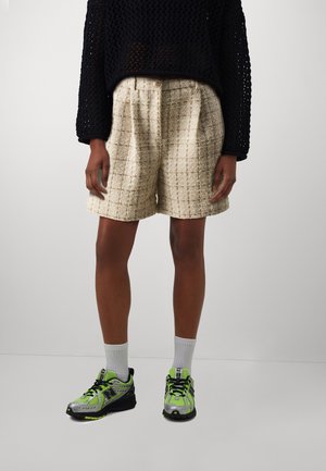 Shorts texturés beige avec un motif à carreaux, associés à un pull en filet noir et des baskets grises avec des accents vert fluo et des chaussettes blanches.