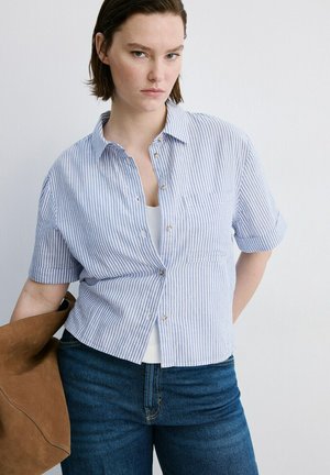 Femme portant une chemise à manches courtes à rayures bleues et blanches, tenant un sac en daim marron, debout devant un fond blanc uni.