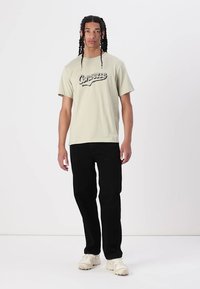 Camiseta de algodón verde claro con el logo de "Converse", combinada con pantalones negros y zapatillas de color crema. Modelo de pie contra un fondo liso.