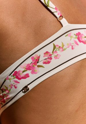 Fehér bikini pánt rózsaszín virágmintával és egy kis fém logó részlettel barnás bőrön, közelkép a vállról és a felső mellkas területéről.