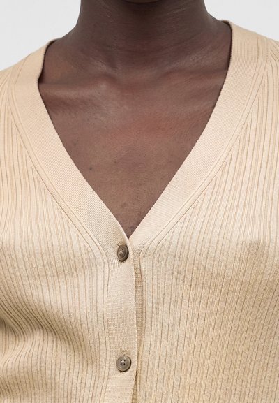 Άτομο με σκούρο δέρμα που φοράει μπεζ ριμπ πλεκτή ζακέτα με δύο κουμπιά κουμπωμένα πάνω από το στήθος και την περιοχή της κλείδας.