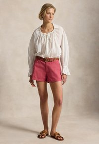 Ξανθιά γυναίκα που φορά λευκό μπλουζάκι με μακριά μανίκια και σούρες στο λαιμό, ροζ σορτς με καφέ ζώνη και καφέ επίπεδα σανδάλια, στέκεται χαλαρή.