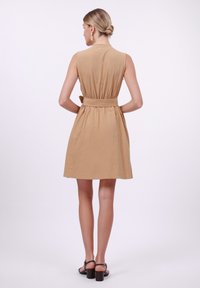 Tan sleeveless dress with a belted waist, textured fabric, and a flared skirt. Styled with black block-heeled sandals.