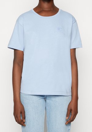 Personne portant un t-shirt Tommy Hilfiger bleu clair à manches courtes et un jean bleu clair, visible du cou aux hanches sur un fond uni.