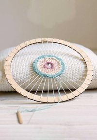 Circular wooden loom with light brown pegs, featuring pastel yarn in a concentric design of pink and blue. Set on a textured white fabric surface.