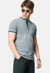 Szara koszulka polo z zielonym kołnierzykiem i akcentami na rękawach, standardowy krój, miękka tkanina, z zapięciem na dwa guziki, noszona z ciemnymi spodniami i okularami przeciwsłonecznymi.