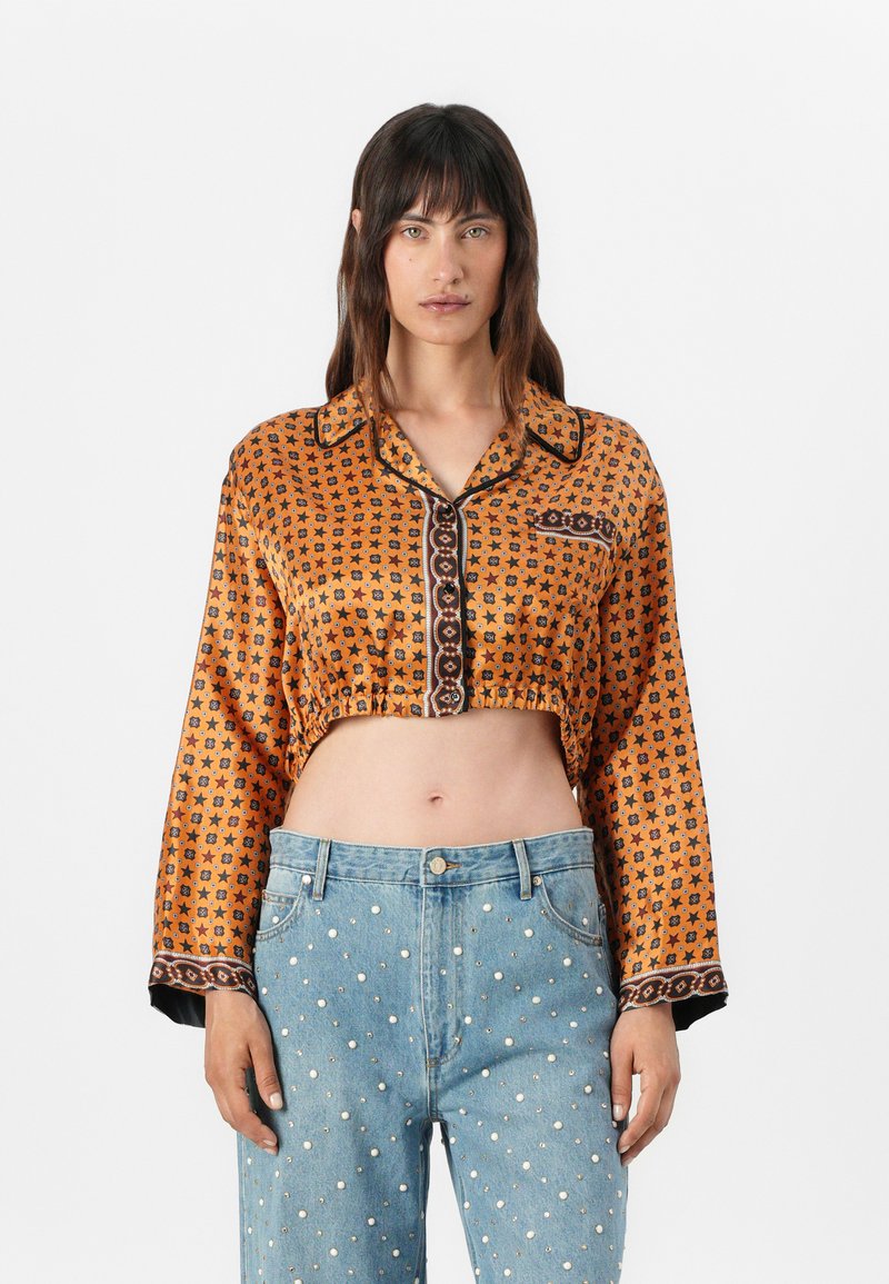 Taillierte orangefarbene Bluse mit Sternenmuster, schwarzen Akzenten und glänzendem Knopfverschluss; kombiniert mit hellblauen Denim-Jeans mit Perlendetails.