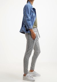Jeansjacka, olivgrön skjorta, grå avsmalnande mjukisbyxor med svarta sidopaneler, vita sneakers. Modellen står framför en enfärgad bakgrund.