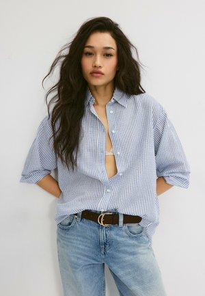 Junge Frau mit langen dunklen Haaren, trägt ein blau-weiß gestreiftes Hemd, das über einem BH aufgeknöpft ist, in hellblaue Jeans mit schwarzem Gürtel gesteckt.