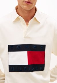 Μπλούζα πόλο εκρού με εξέχον λογότυπο σε μπλε ναυτικό, κόκκινο και λευκό, με κεντητό κείμενο "TOMMY HILFIGER." Λεπτομέρεια γιακά με κουμπιά.