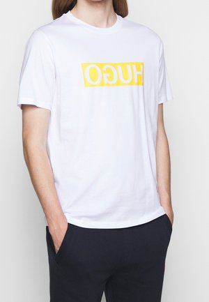 Osoba ubrana w białą koszulkę z krótkim rękawem, na której znajduje się żółty prostokątny logo z odwróconym napisem "HUGO", oraz czarne spodnie, z rękami w kieszeniach.