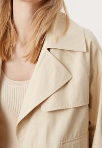 Beżowy bawełniany płaszcz trencz z szerokim kołnierzem i kieszenią na przodzie, noszony na lekkiej, prążkowanej bluzce bez rękawów, eksponujący gładkie faktury.