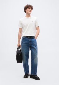 Νεαρός άνδρας με λευκό t-shirt Calvin Klein, μπλε τζιν, μαύρη ζώνη, μαύρα παπούτσια, κρατώντας μαύρη τσάντα χειρός, στέκεται μπροστά σε μονόχρωμο φόντο.