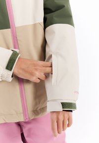 Veste en blocs de couleurs beige, verte et crème avec une fermeture éclair rose, des poignets élastiques et une poche interne accessible par une ouverture dans la manche.
