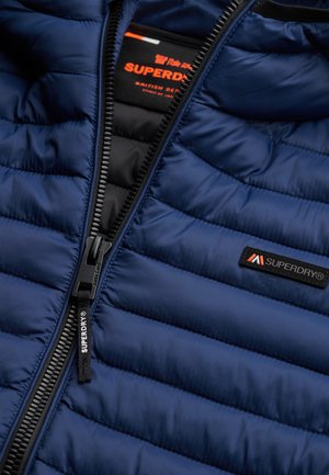 Chaqueta acolchada azul marino con cremallera negra, logotipo de Superdry en el pecho y en el tirador de la cremallera, parcialmente desabrochada revelando la etiqueta interior.