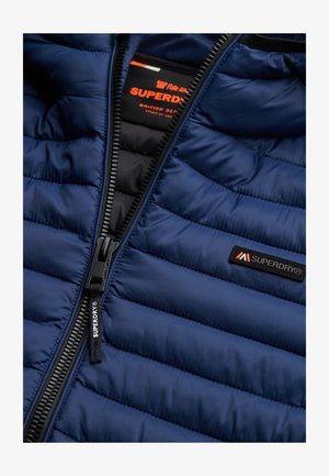 Marineblau gesteppte Jacke mit schwarzfarbenem Reißverschluss, Superdry-Logo auf der Brust und am Reißverschluss, teilweise geöffnet, sodass das Innenetikett sichtbar ist.