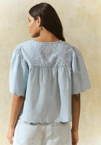 Light blue blouse with short, puffed sleeves, scalloped hem, and textured detailing on the back, made of a soft, woven fabric.