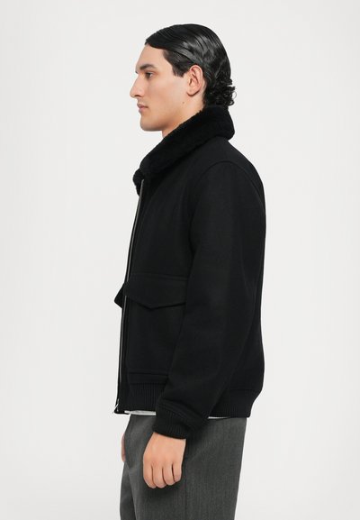 Schwarze Bomberjacke mit einem Kunstpelzkragen, Reißverschluss und einer aufgesetzten Tasche vorne; verfügt über gerippte Bündchen und einen gerippten Saum mit einer glatten Textur.