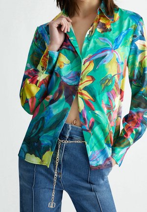 Femme portant un chemisier coloré à imprimé tropical avec des boutons et un jean bleu taille haute accessoirisé d'une ceinture chaîne en or, main dans la poche.