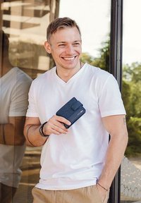 Blue fabric wallet held in a hand, featuring a button closure. Man wears a white V-neck t-shirt and light-colored pants, smiling in front of glass.