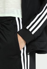 Main dans la poche, portant un pantalon de survêtement noir et une veste avec trois bandes blanches, ainsi qu'une chemise légère translucide visible au-dessus de la taille.