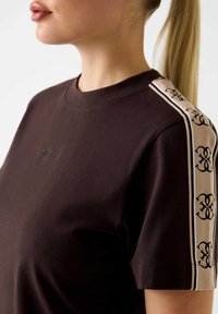 Brązowa bawełniana koszulka z krótkim rękawem, z dekoltem w serek i logo na klatce piersiowej. Boki mają beżowy kolor z czarnymi wzorzystymi akcentami.