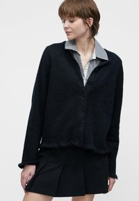 Mladá žena oblečená v černém cardigan s knoflíky přes kostkovanou košili a černou plisovanou sukni, stojící na pozadí bílé stěny.