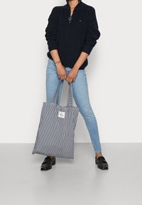 Sac fourre-tout à rayures verticales noires et blanches, design spacieux, poignées robustes et étiquette de logo visible. Porté avec un pull zippé et un jean.