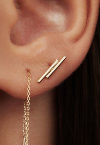 Gold-tone earrings with a minimalist design featuring two vertical bars and a delicate chain hanging from one, set against an ear.