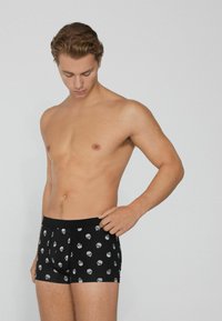 Mladý muž se dívá dolů, oblečený v černých boxerkách s bílým vzorem lebek na čistém světlém pozadí.