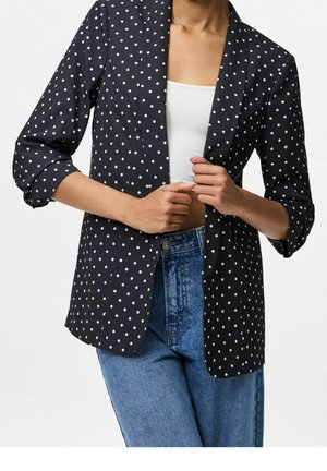 Personne portant un blazer noir à pois blancs, un top court blanc et un jean bleu taille haute, tenant les revers du blazer.