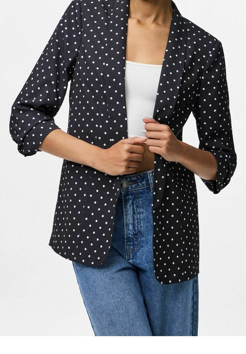 Personne portant un blazer noir à pois blancs, un top court blanc et un jean bleu taille haute, tenant les revers du blazer.