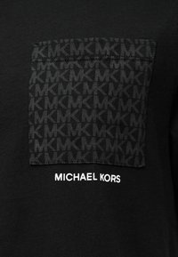 Μαύρο μπλουζάκι με υφασμάτινο έμπλαστρο με επαναλαμβανόμενο μοτίβο MK και την επιγραφή "MICHAEL KORS" σε λευκό κάτω από την τσέπη.