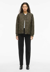 Prešita bombažna jakna olivno zelene barve z zadrgo spredaj, rebrastimi manšetami in stranskimi žepi, nošena preko svetlega topa in črnih hlač.