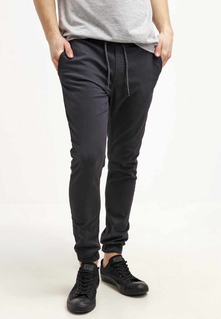 Tmavě šedé teplákové kalhoty z hladké látky, s zužujícími se nohavicemi, elastickými lemy a stahovací šňůrkou v pase. Nošeny s černými teniskami.