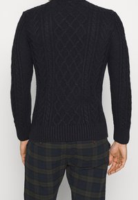 Pull bleu marine tricoté avec motifs en torsades, poignets et ourlet côtelés. Porté avec un pantalon écossais foncé. Texture détaillée visible.