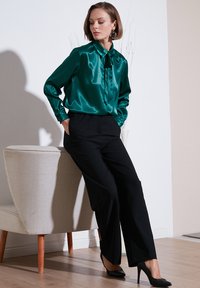 Teal satin blouse with a bow tie detail, paired with high-waisted black trousers and black stilettos, set against a light background.