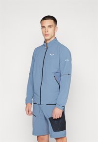Lyseblå treningsjakke med høy krage, svart glidelås og logo; kombinert med matchende shorts med en svart nettlomme.