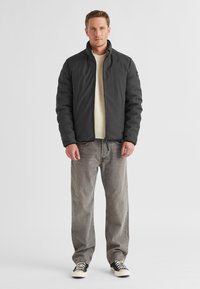 Dunkelgraue Steppjacke mit hohem Kragen, getragen über einem cremefarbenen Strickpullover. Kombiniert mit hellgrauen Jeans und schwarzen Sneakers mit weißen Akzenten.