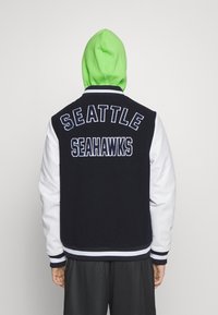 Schwarz-weiße Collegejacke mit aufgesticktem "SEATTLE SEAHAWKS" auf dem Rücken, ergänzt durch einen grünen Kapuzenpullover, der darunter hervorblitzt.