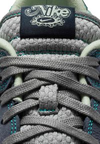 Lähikuva harmaan kudotun Nike-tennarikielen ja nauhojen teal-ompeleista sekä laivastonsinisestä merkistä, jossa on Nike-logo.