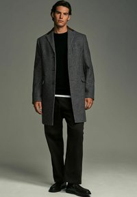 Grå houndstooth frakke med en brun krave, dobbeltknaplukning, to sidelommer, båret over en sort sweater og sorte bukser.