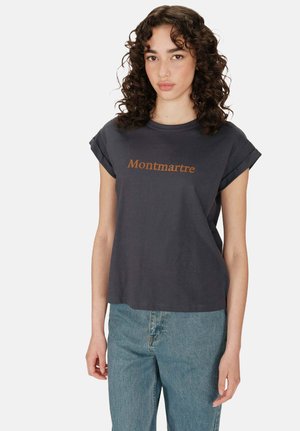 T-shirt grigio scuro con maniche arrotolate e la scritta "Montmartre" stampata in arancione, realizzato in tessuto di cotone. Abbinato a jeans blu chiaro.